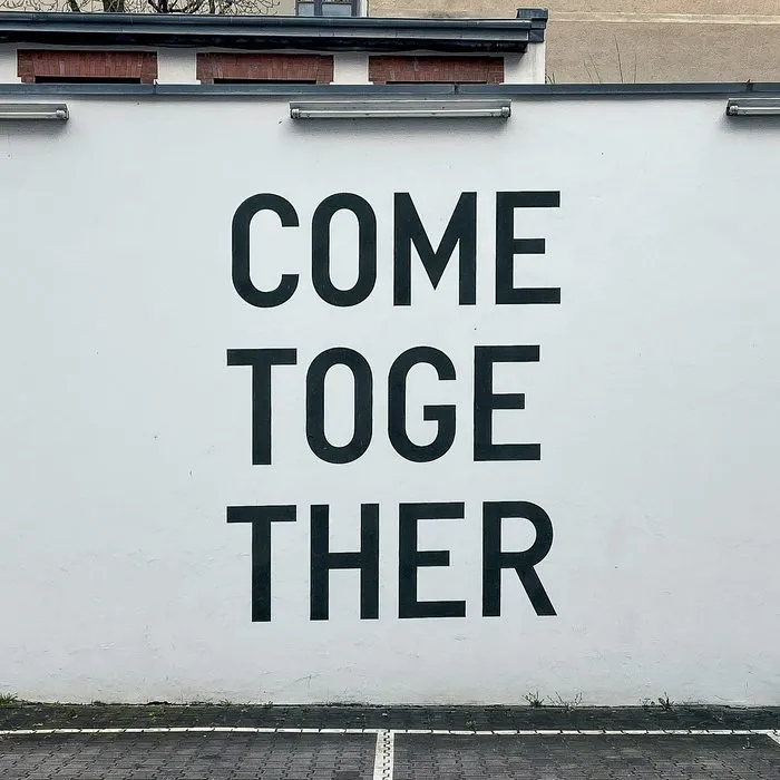 Pared blanca con las palabras «Come together» en grandes letras negras